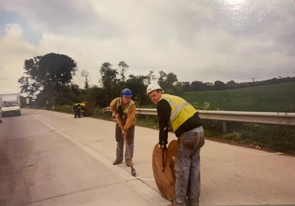 Two workers finishing a fresh concrete road surface with a rake and a bull float, paving machine behind