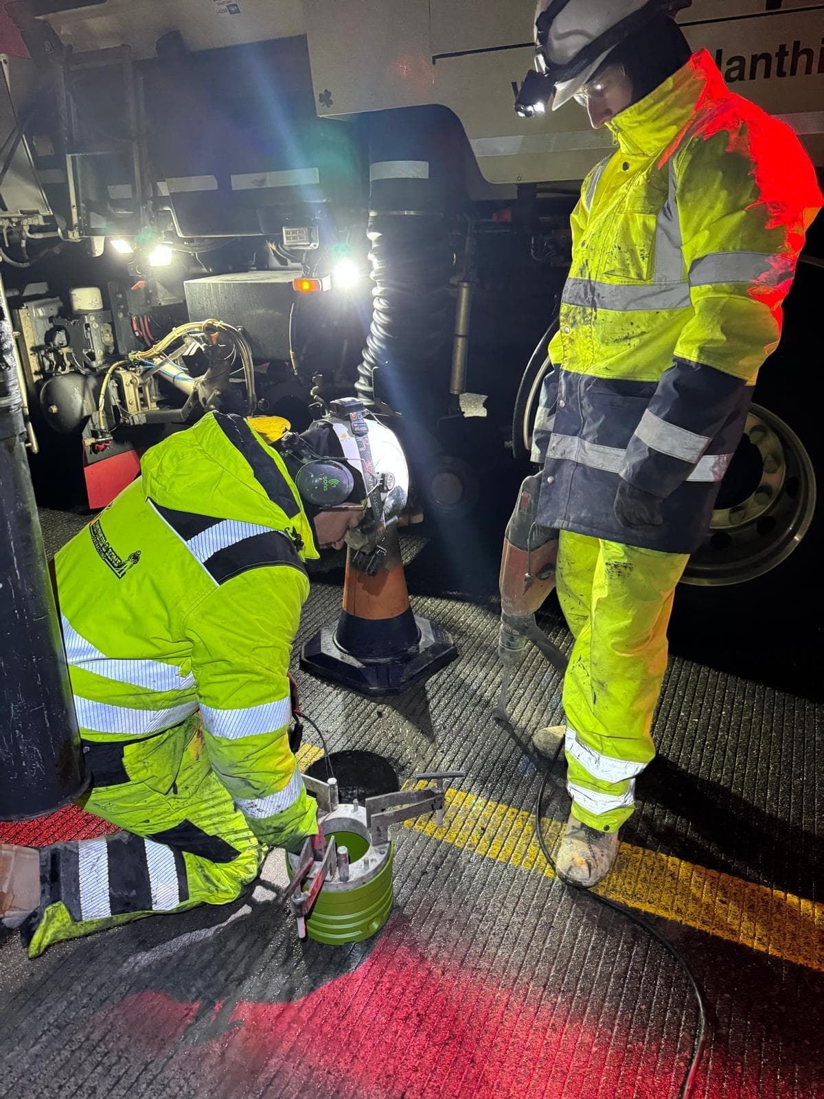 Two workers in yellow hi-vis at night working at the rear of a machine lit by work lights