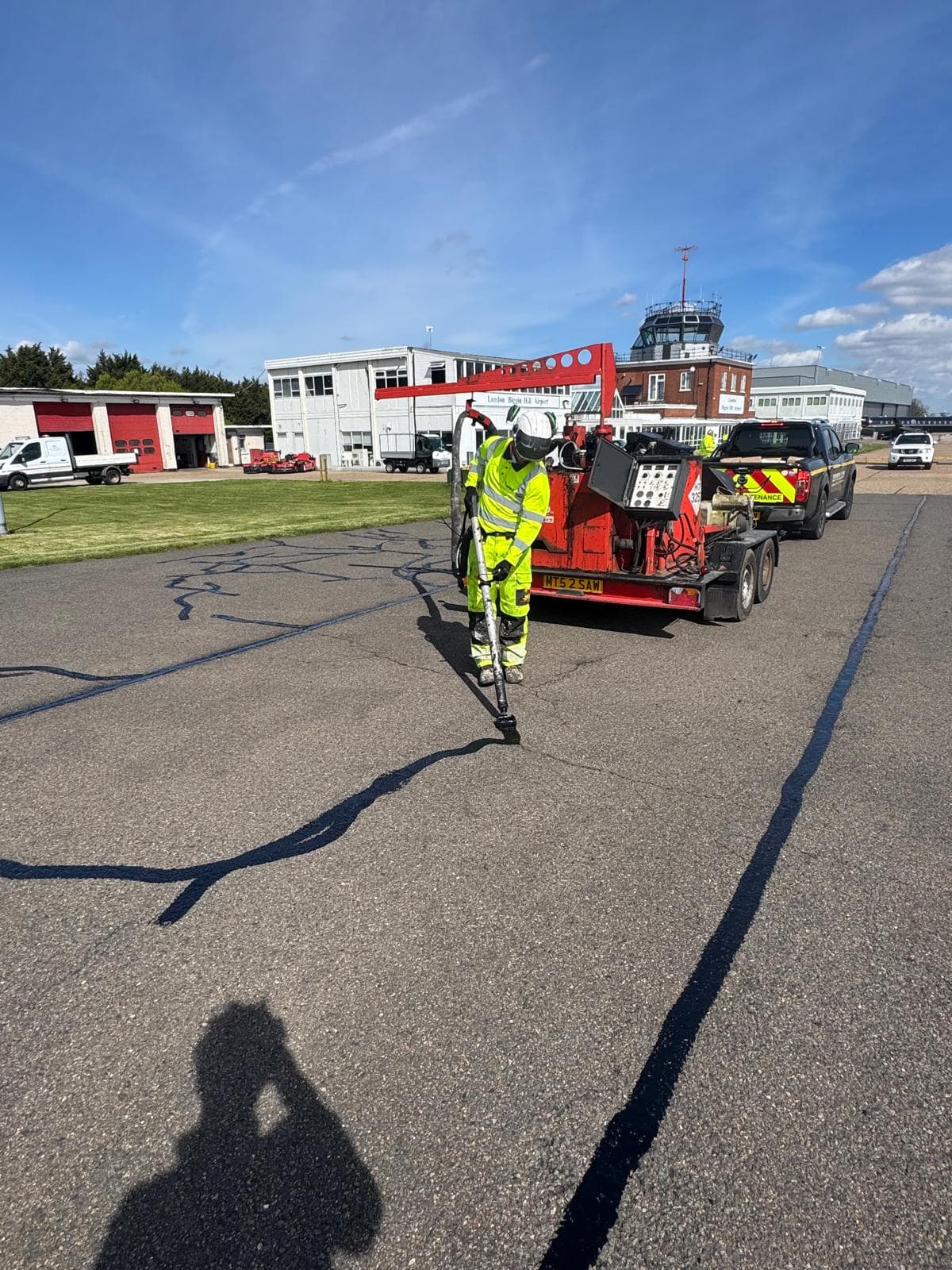 Worker in hi-vis applying crack-sealant on an airfield apron with a red trailer and branded Shepherd and Sons pickup