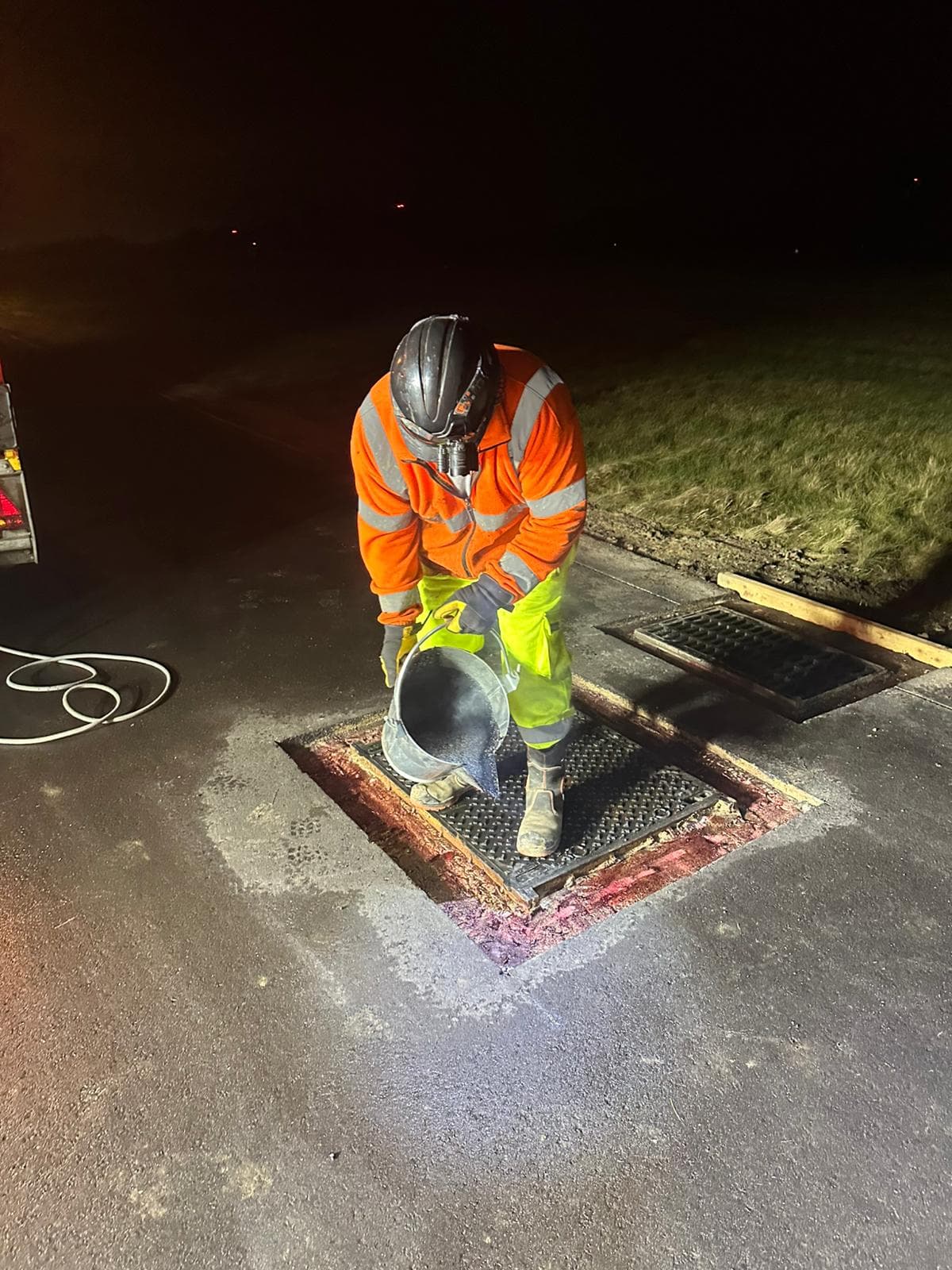 Operative in hi-vis pouring material from a bucket into an open metal drainage grate at night